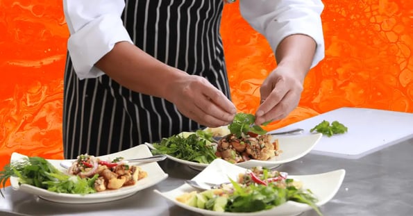 The bottom half of a man’s body wearing a white long-sleeved shirt and a black-and-white pinstripe apron assembling plates of food.